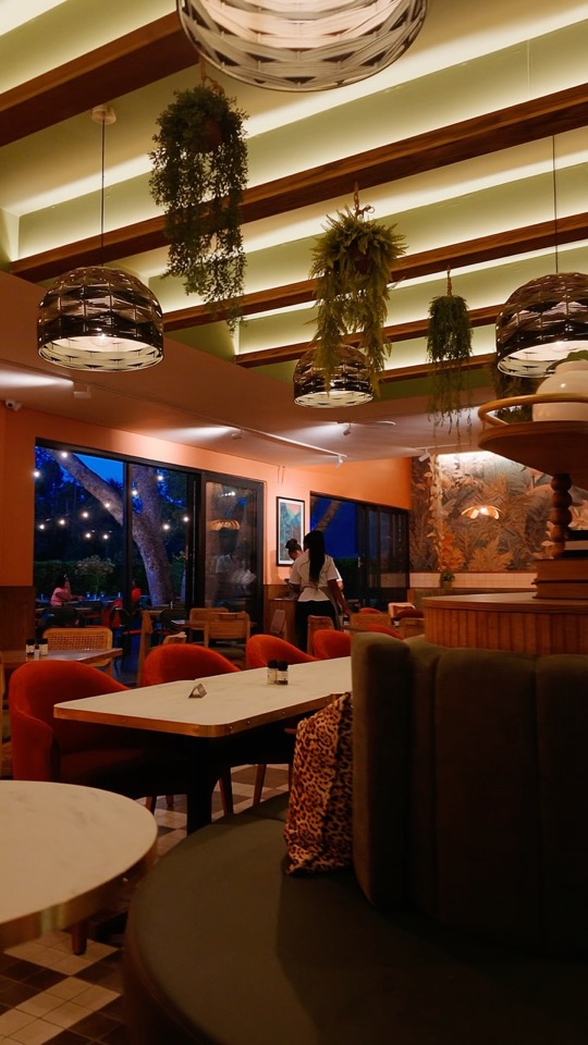 The dining room at dusk: orange velvet chairs, marble tables, hanging plants under woven pendant lights and a garden with string lights beyond the windows